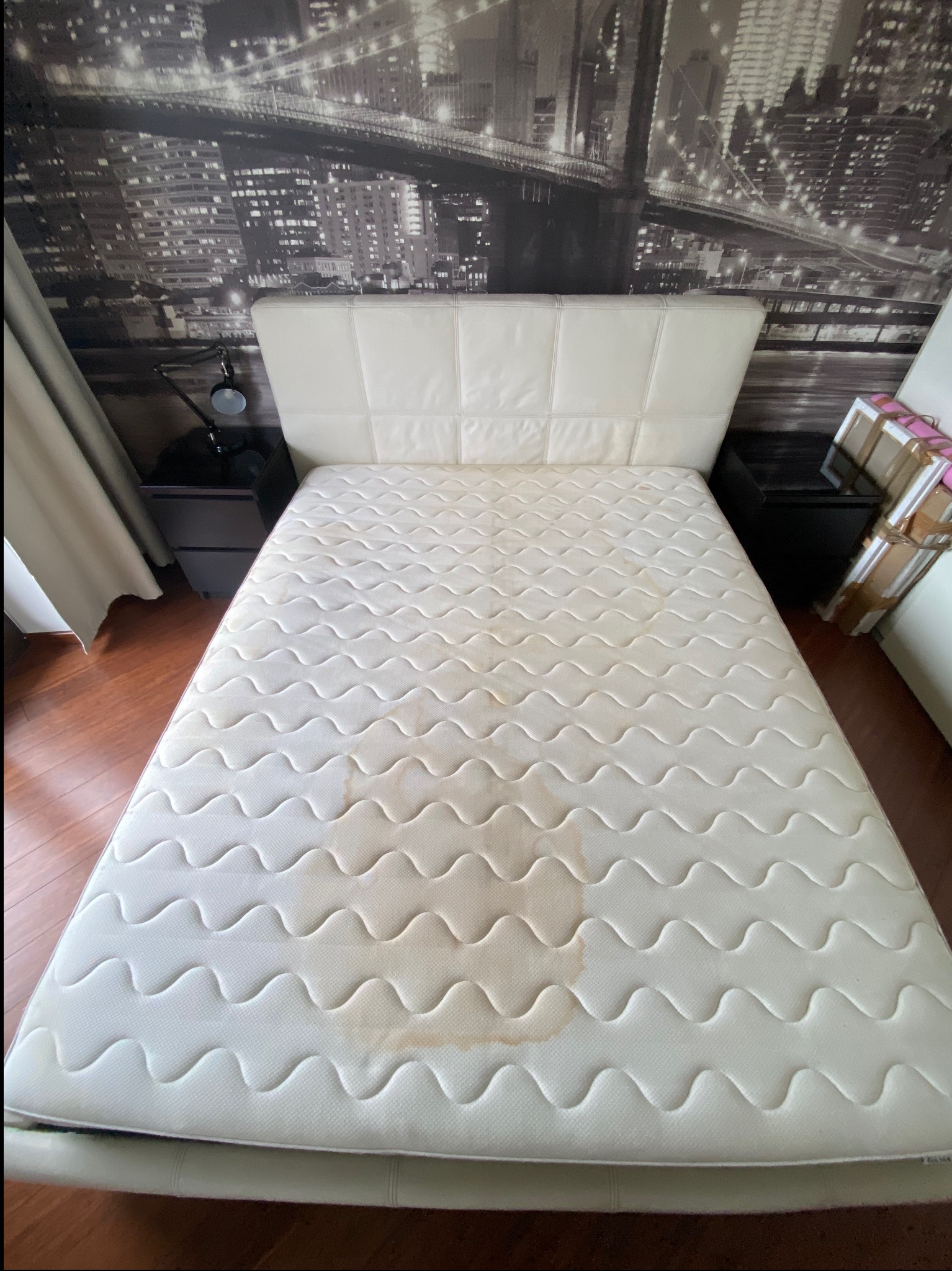 Zabrudzony materac na łóżku w Sułkowicach, widoczne plamy. Tapeta z motywem miasta w tle. Materac wymaga odświeżenia i profesjonalnego czyszczenia.