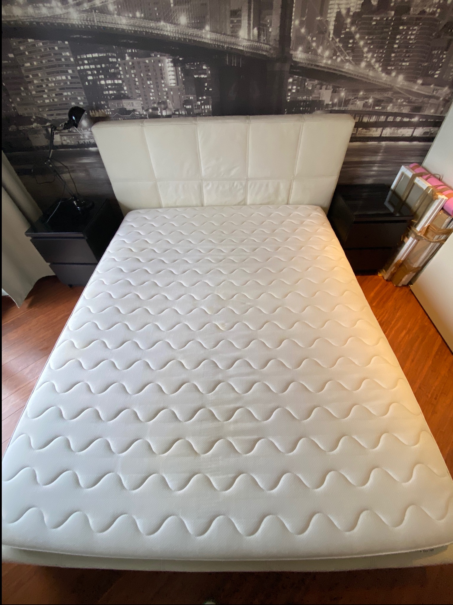 Biały materac na łóżku z pikowanym zagłówkiem, widok z góry. W tle nocny pejzaż miasta na fototapecie i czarne szafki nocne z lampką.