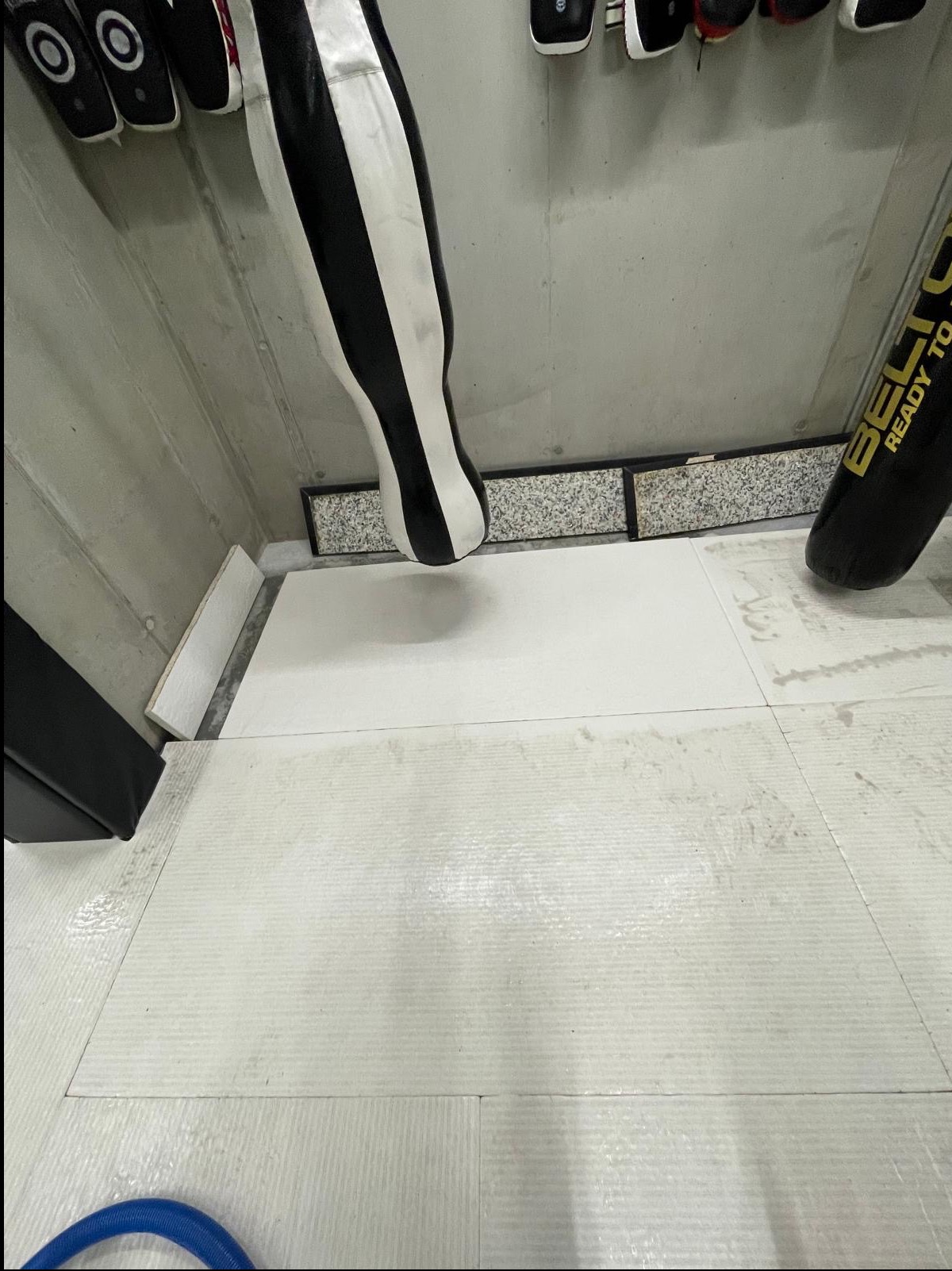 Biała podłoga z piankowych puzzli po czyszczeniu, widoczne ślady wilgoci i fragment węża odkurzacza wodnego w rogu. Sprzęt do sportów walki przy ścianie.