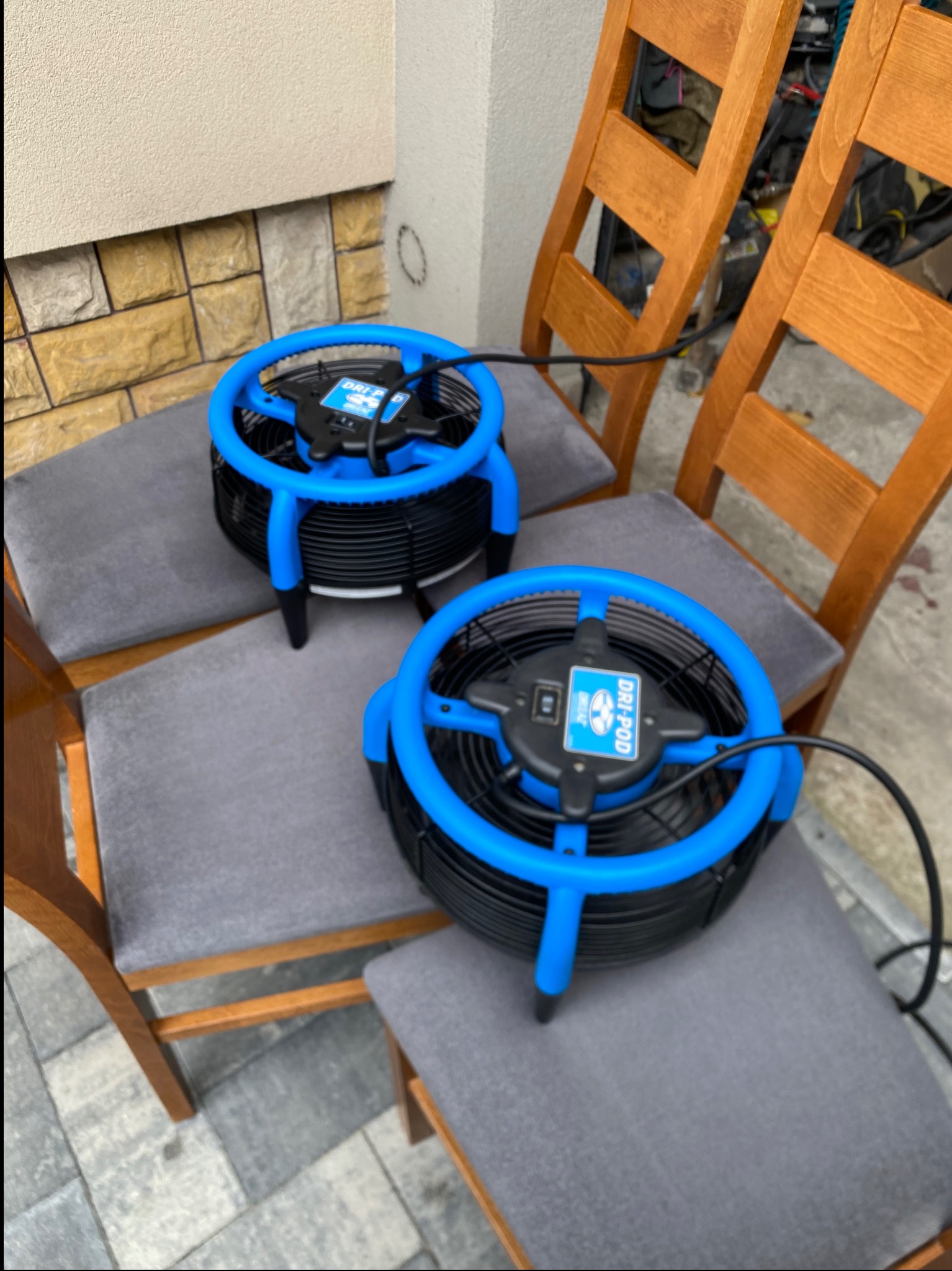 Dwa wentylatory osuszające Dri-Pod na krzesłach z szarą tapicerką, ustawione na zewnątrz budynku z beżową elewacją i kostką brukową. Widoczne kable zasilające.