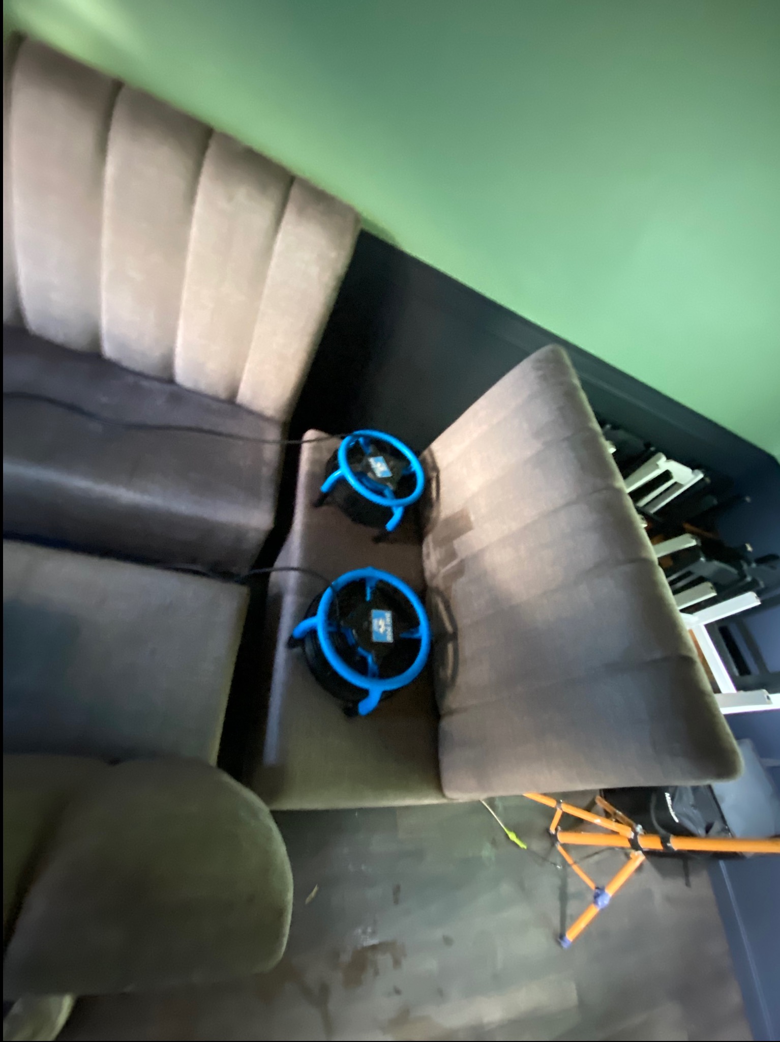 Dwa wentylatory z niebieskimi obudowami suszą tapicerkę sofy w odcieniach beżu, po zabiegu czyszczenia. Widoczny fragment podłogi i ściany.