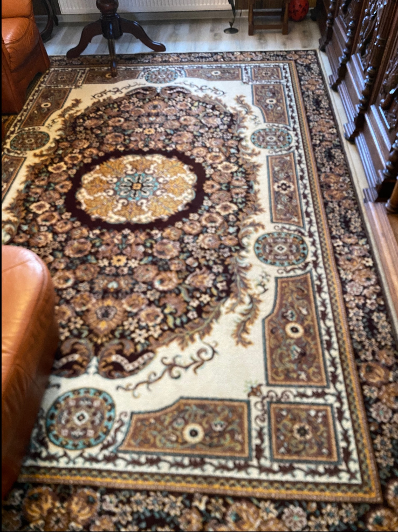 Wzorzysty dywan orientalny z motywem kwiatowym, leżący na podłodze z paneli, częściowo zasłonięty przez meble. Widoczne bogate zdobienia i detale.