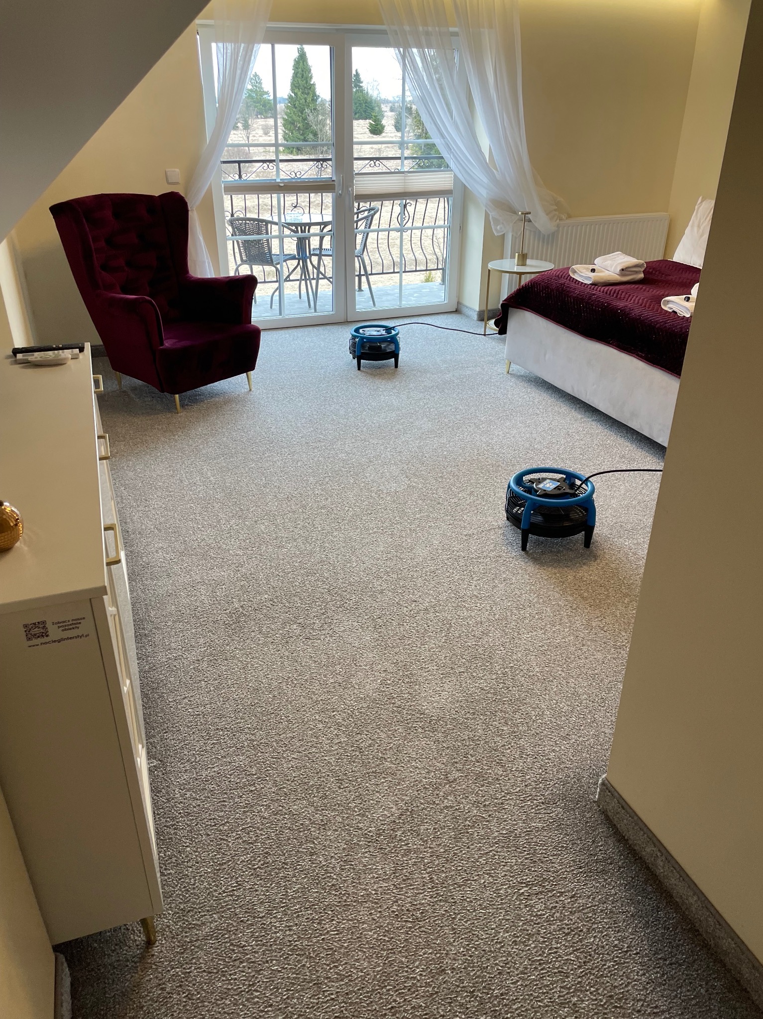 Pokój hotelowy z szarym dywanem suszonym przez dwa niebieskie wentylatory po czyszczeniu. W tle balkon z widokiem na góry, fotel i łóżko.