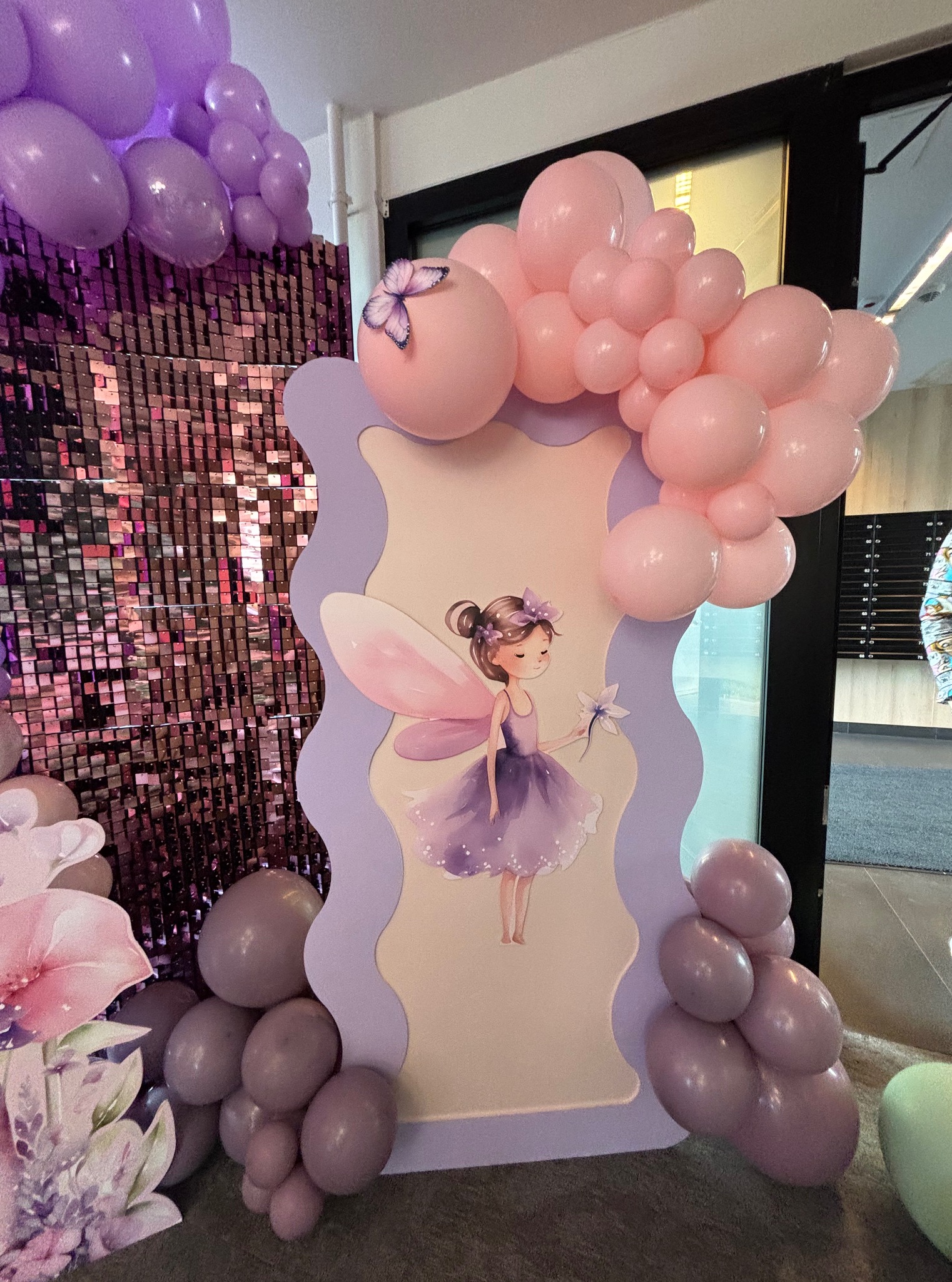 Dekoracja urodzinowa: postać wróżki w fioletowej ramce, otoczona różowymi i fioletowymi balonami na tle błyszczącej ściany z cekinów.