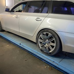 Auto-Pablo - Srebrne kombi BMW na podnośniku warsztatowym z urządzeniem do geometrii kół przy przednim kole. Widok z boku, oświetlenie warsztatowe.