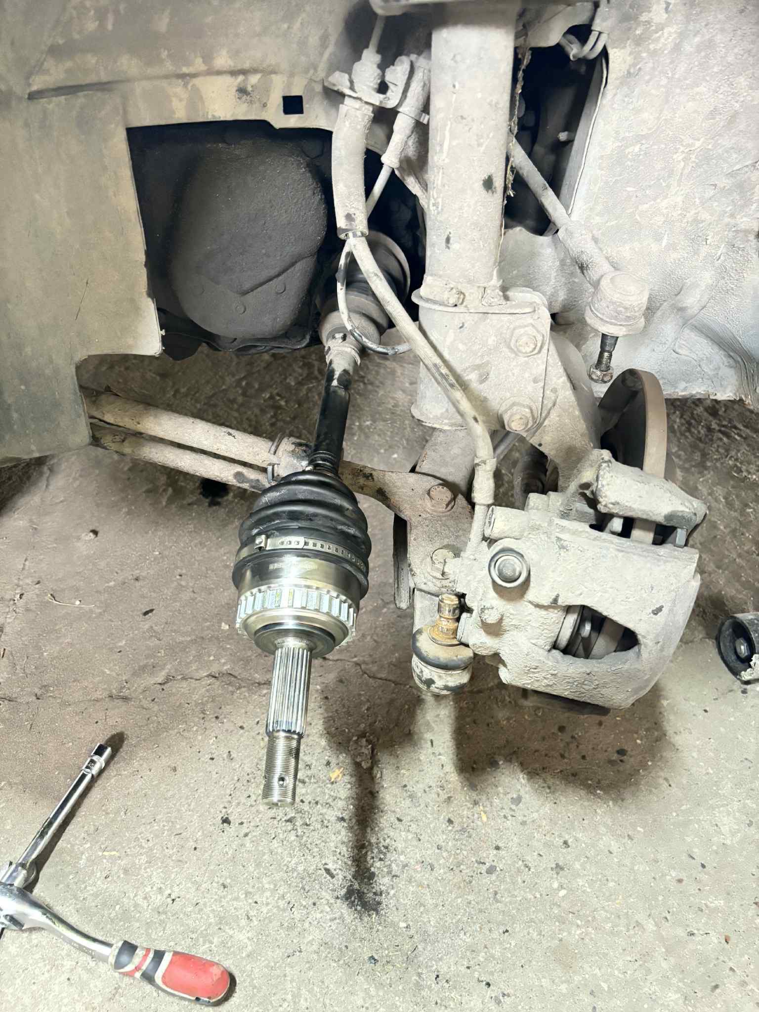 Wymontowana półoś napędowa z przegubem, widoczne elementy zawieszenia i hamulca tarczowego. Narzędzia warsztatowe na podłożu. Auto w trakcie naprawy.