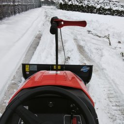 Pogotowie Ogrodowe Daniel Janowiec - Czerwony traktor z pługiem odśnieża zaśnieżoną drogę w pochmurny dzień. Widok z perspektywy operatora, ślady opon w śniegu. W tle domy i drzewa pod śniegiem.