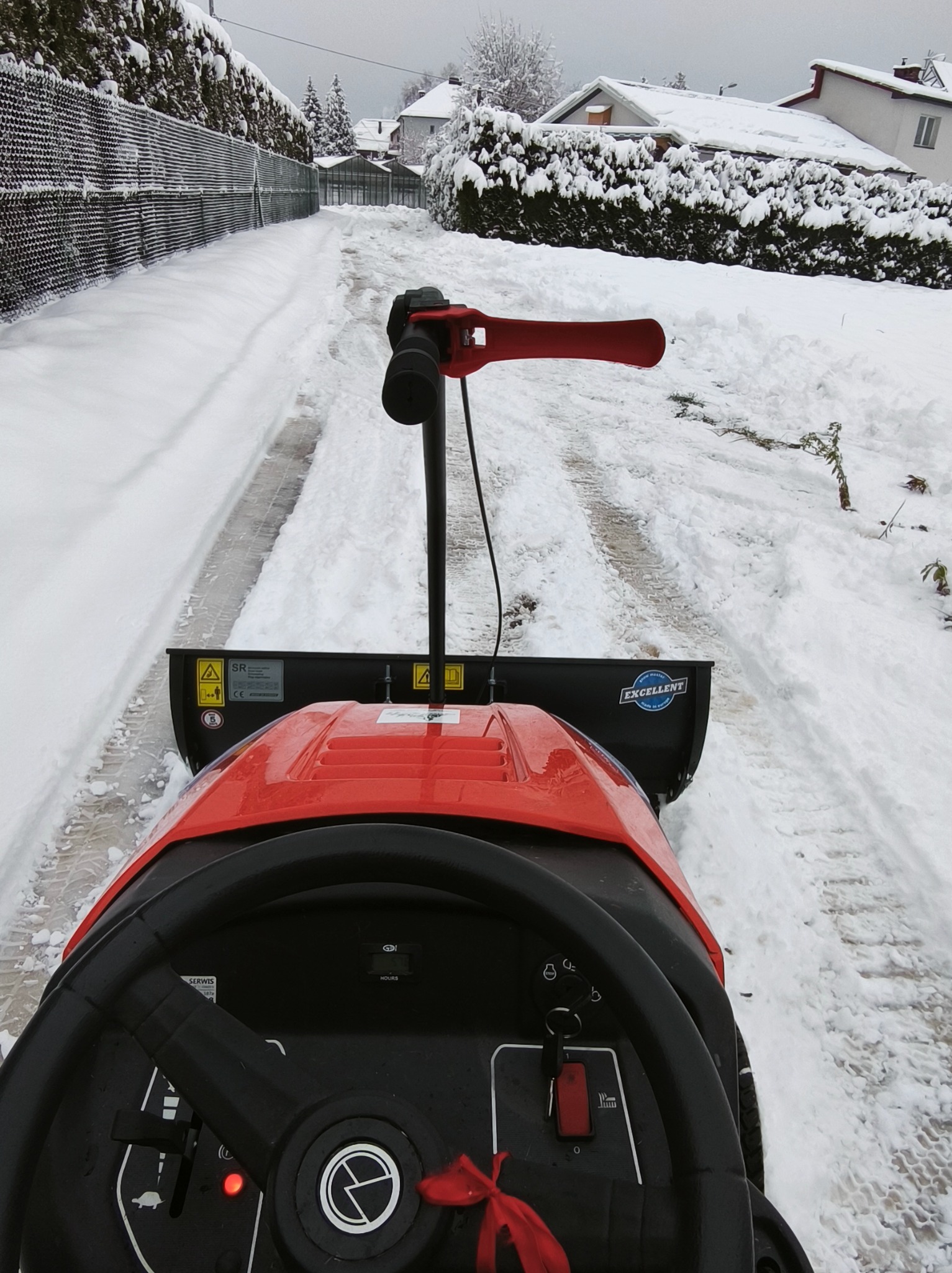 Czerwony traktor z pługiem odśnieża zaśnieżoną drogę w pochmurny dzień. Widok z perspektywy operatora, ślady opon w śniegu. W tle domy i drzewa pod śniegiem.