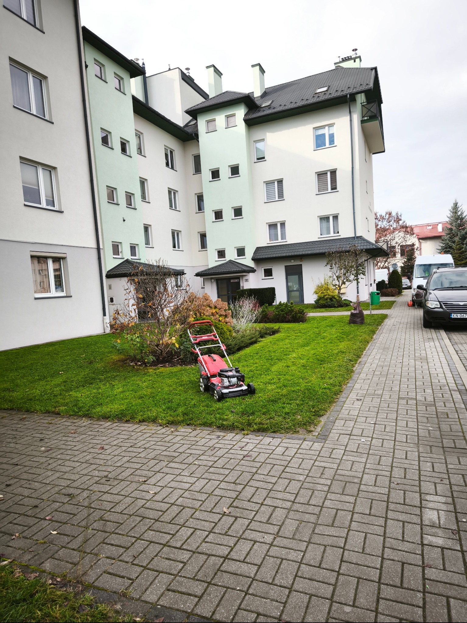 Czerwona kosiarka spalinowa na trawniku przed nowoczesnym budynkiem mieszkalnym. Widok z poziomu chodnika z kostki brukowej, pochmurne niebo w tle.