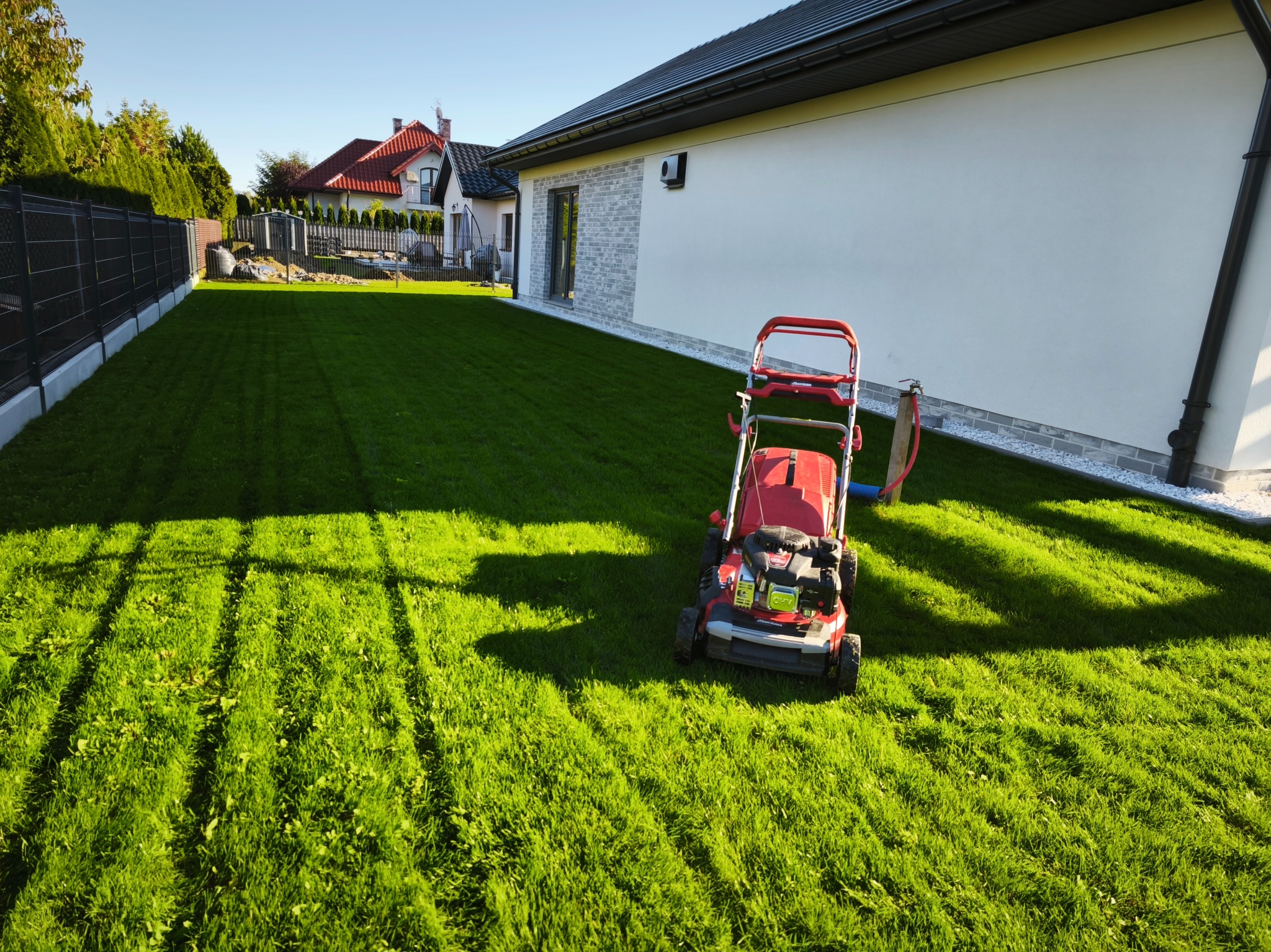 Czerwona kosiarka na tle świeżo skoszonego, zielonego trawnika przed nowoczesnym domem z białymi ścianami i szarym dachem w słoneczny dzień. Widoczne pasy po koszeniu.