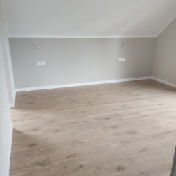 MEMKO - Jasne panele podłogowe w nowym pokoju z beżowymi ścianami, widoczne białe listwy przypodłogowe i gniazdka elektryczne. Ukończone wykończenie wnętrza.