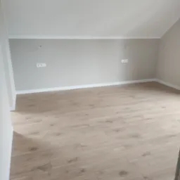 Jasne panele podłogowe w nowym pokoju z beżowymi ścianami, widoczne białe listwy przypodłogowe i gniazdka elektryczne. Ukończone wykończenie wnętrza.