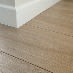 MEMKO - Jasne panele podłogowe ułożone przy białej ścianie z przypodłogową listwą. Widoczne łączenia paneli i subtelna faktura drewna.