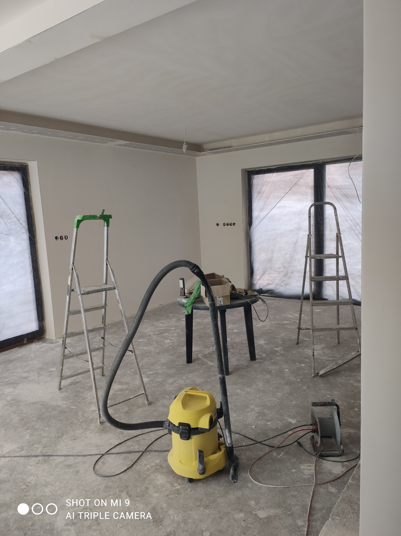 Wnętrze w trakcie wykańczania: świeżo wygładzone ściany, drabiny, odkurzacz przemysłowy i stół z narzędziami. Okna zabezpieczone folią.