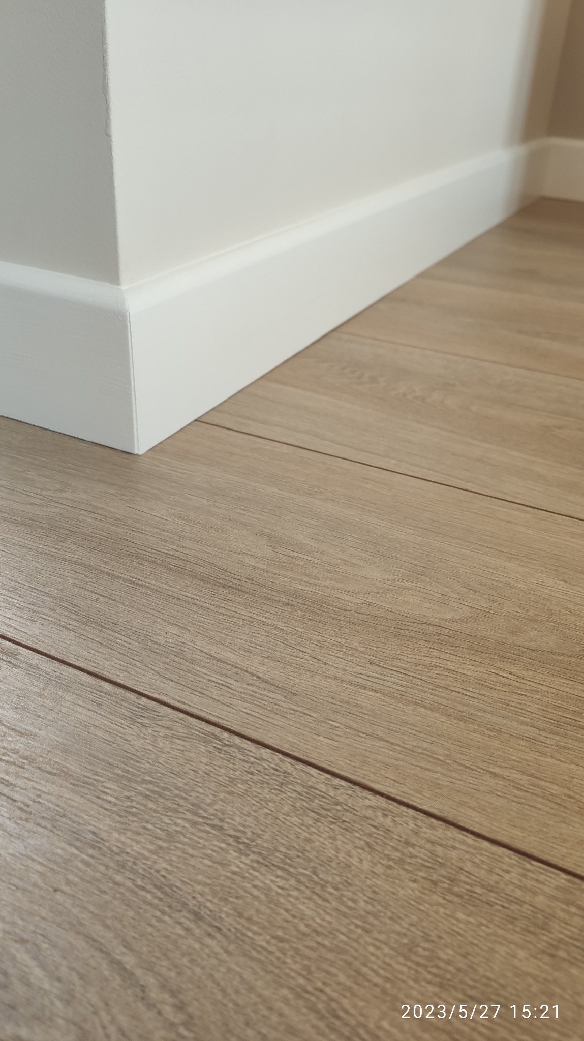 Jasne panele podłogowe ułożone przy białej ścianie z przypodłogową listwą. Widoczne łączenia paneli i subtelna faktura drewna.