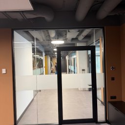 FIRMA BUDOWLANA OLEKSANDR BOICHENKO - Nowoczesne biuro z przeszklonymi ścianami i drzwiami z matowym pasem. Widoczne instalacje pod sufitem i osoba na drabinie w głębi. Minimalistyczny design.