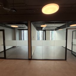 FIRMA BUDOWLANA OLEKSANDR BOICHENKO - Nowoczesne biuro po malowaniu: przeszklone ściany działowe z matową folią, brązowa podłoga, widoczne lampy sufitowe. Minimalistyczny design, czyste linie, jasne wnętrze.