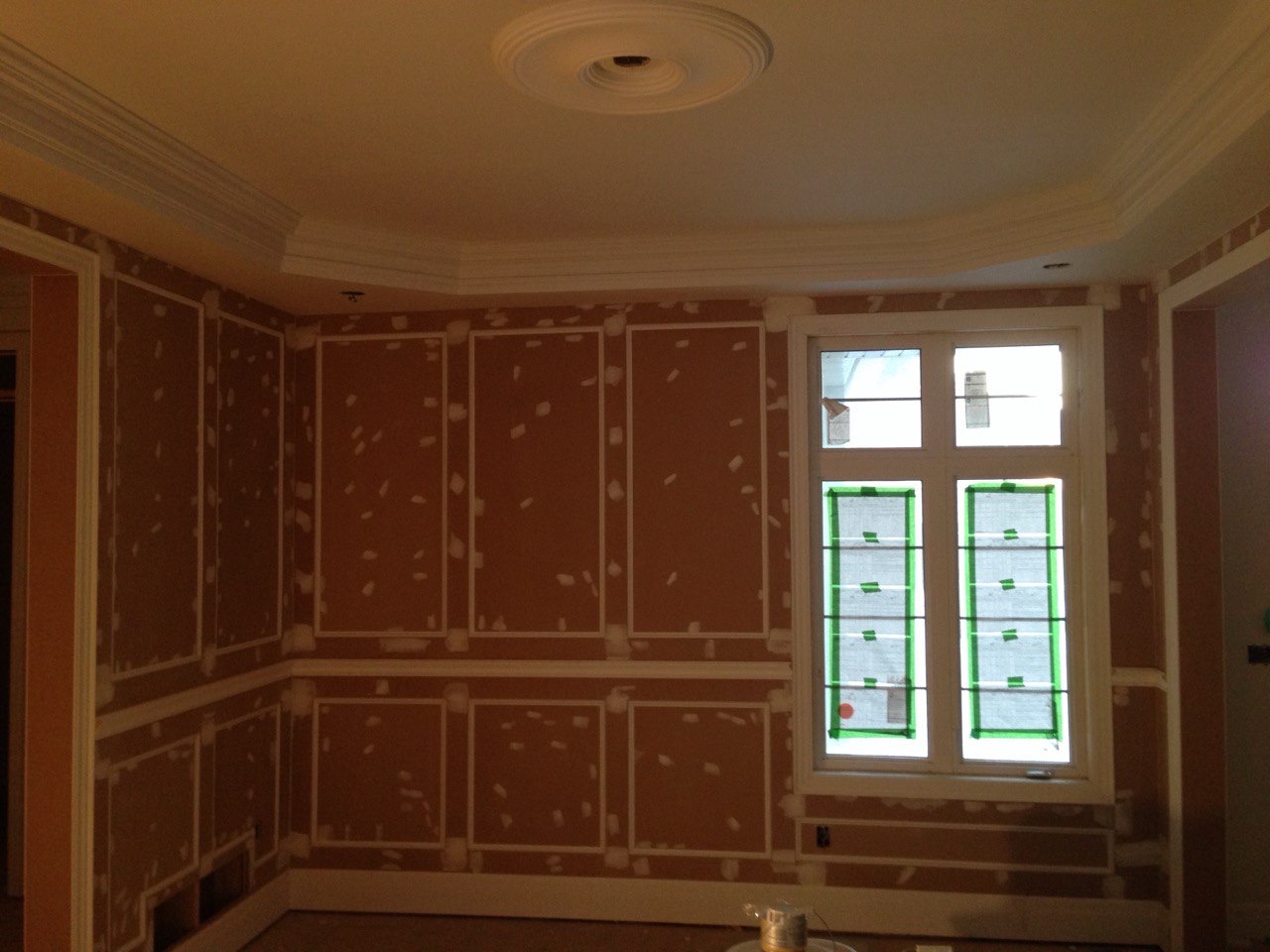 Pokój w trakcie szpachlowania z dekoracyjnymi listwami i zabezpieczonym oknem. Widoczne ślady szpachli na ścianach i suficie, przygotowanie do malowania.