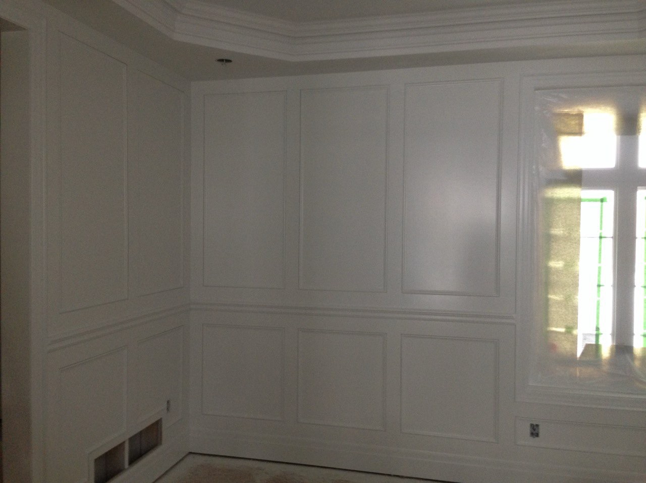 Białe panele ścienne z listwami w klasycznym stylu, okno zabezpieczone folią malarską, widok na wykończone wnętrze z detalami sztukaterii.