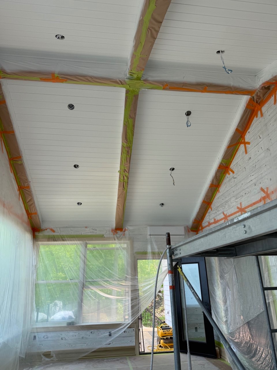 Białe, drewniane panele sufitowe z taśmą malarską, zabezpieczone folią okna i ściany, widok z rusztowania. Przygotowanie do malowania.