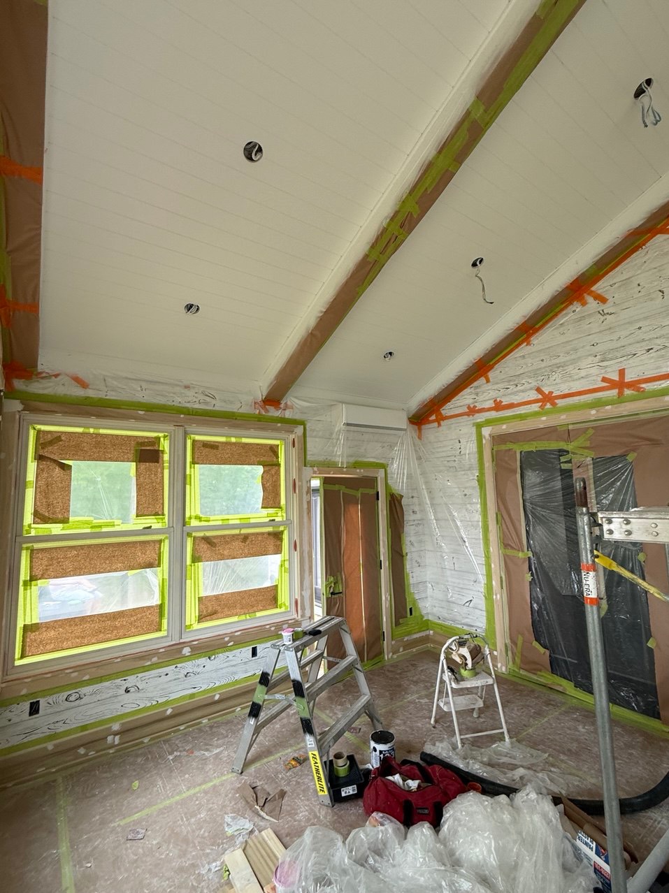 Białe wnętrze ze skośnym sufitem w trakcie zabezpieczania przed malowaniem. Okna, drzwi i listwy zabezpieczone taśmą i folią. Widoczne drabiny i akcesoria malarskie.