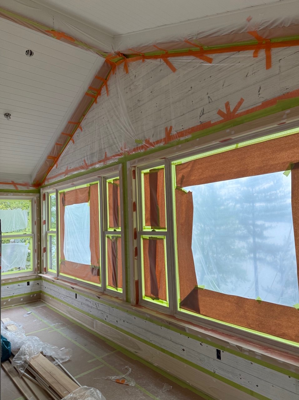 Przygotowane do malowania wnętrze z oknami zabezpieczonymi folią i taśmą malarską. Widoczne drewniane ściany i sufit, również zabezpieczone przed zabrudzeniem.