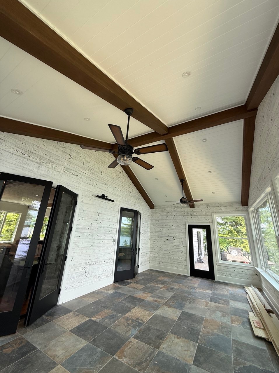 Jasne wnętrze z drewnianą boazerią, brązowymi belkami stropowymi i wentylatorami sufitowymi. Widoczne okna i drzwi z czarnymi ramami.