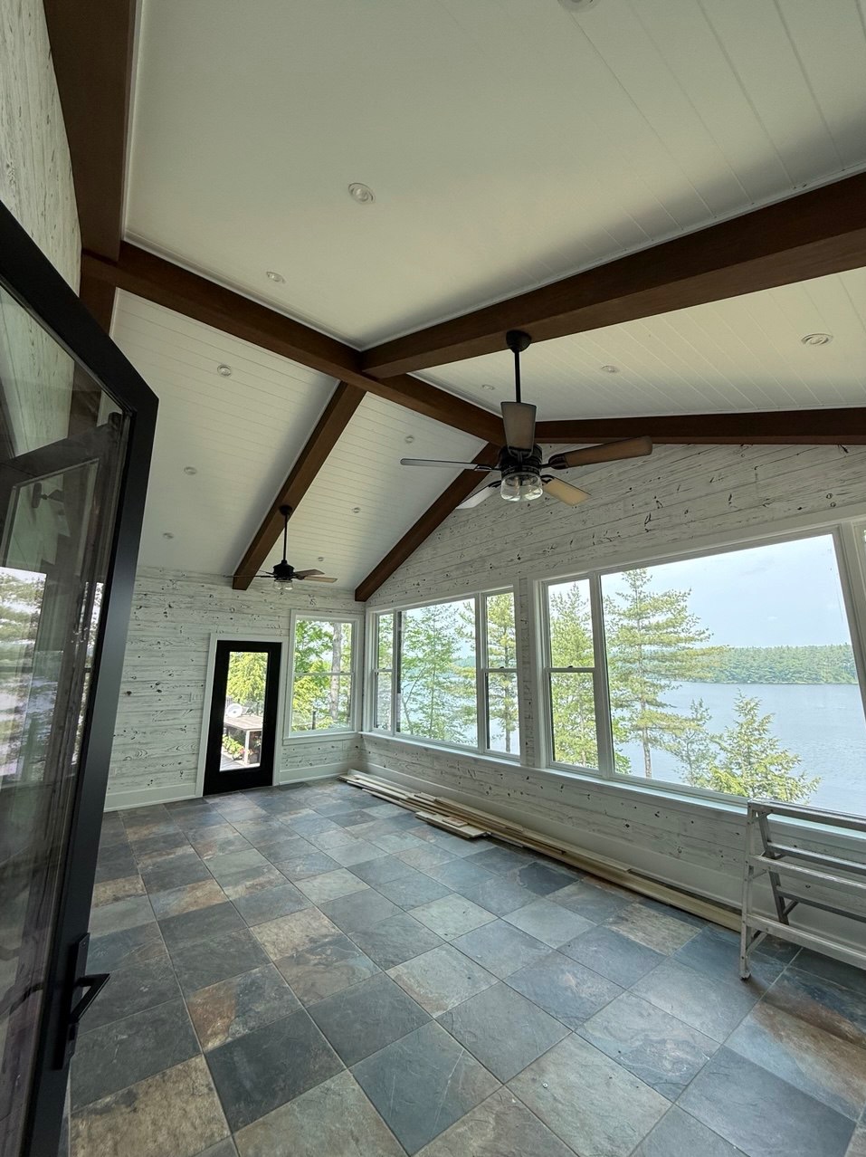 Jasne wnętrze werandy z oknami na jezioro, drewnianą boazerią i stropem z belkami. Widoczne płytki podłogowe i wentylatory sufitowe.