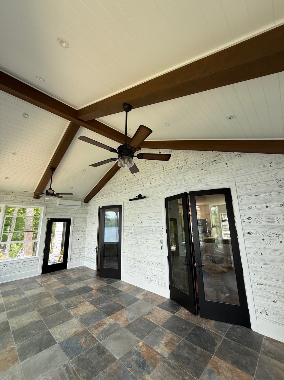 Jasne wnętrze z drewnianymi belkami i panelami ściennymi imitującymi drewno, wentylatory sufitowe i ciemne ramy okienno-drzwiowe. Podłoga wyłożona płytkami.