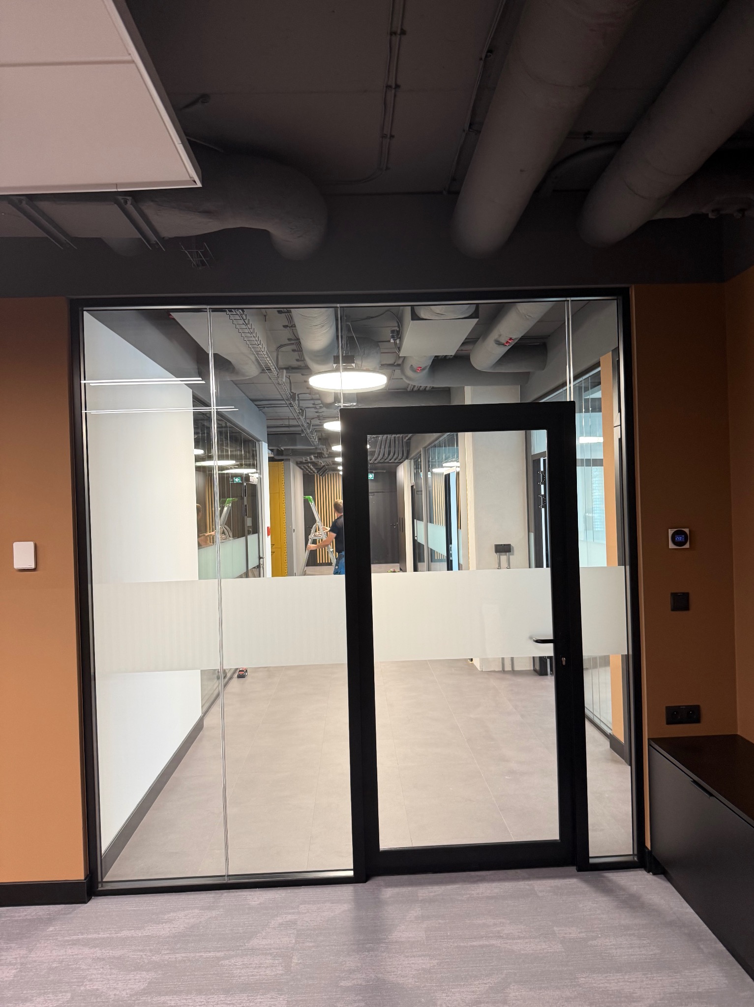 Nowoczesne biuro z przeszklonymi ścianami i drzwiami z matowym pasem. Widoczne instalacje pod sufitem i osoba na drabinie w głębi. Minimalistyczny design.