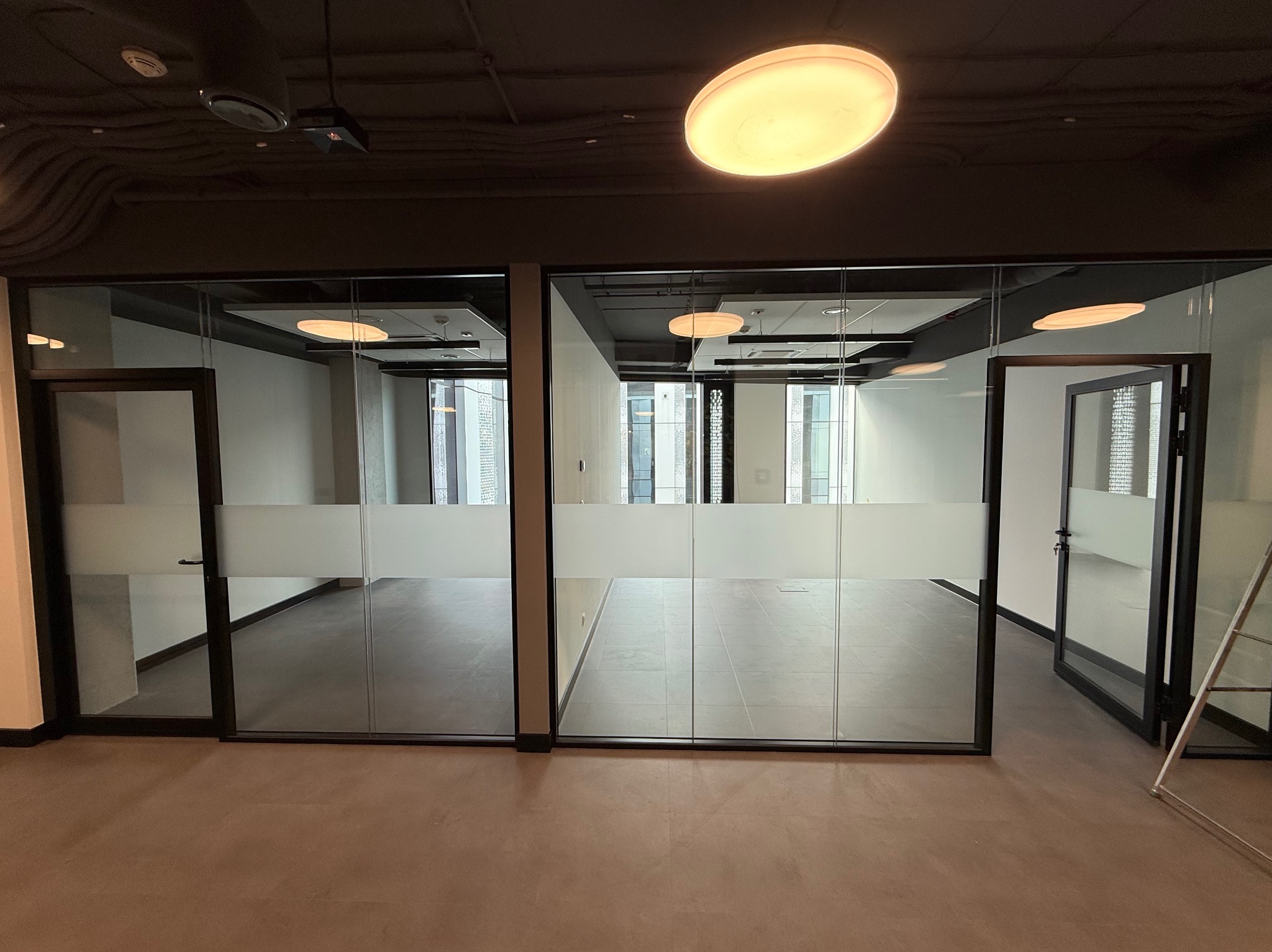 Nowoczesne biuro po malowaniu: przeszklone ściany działowe z matową folią, brązowa podłoga, widoczne lampy sufitowe. Minimalistyczny design, czyste linie, jasne wnętrze.