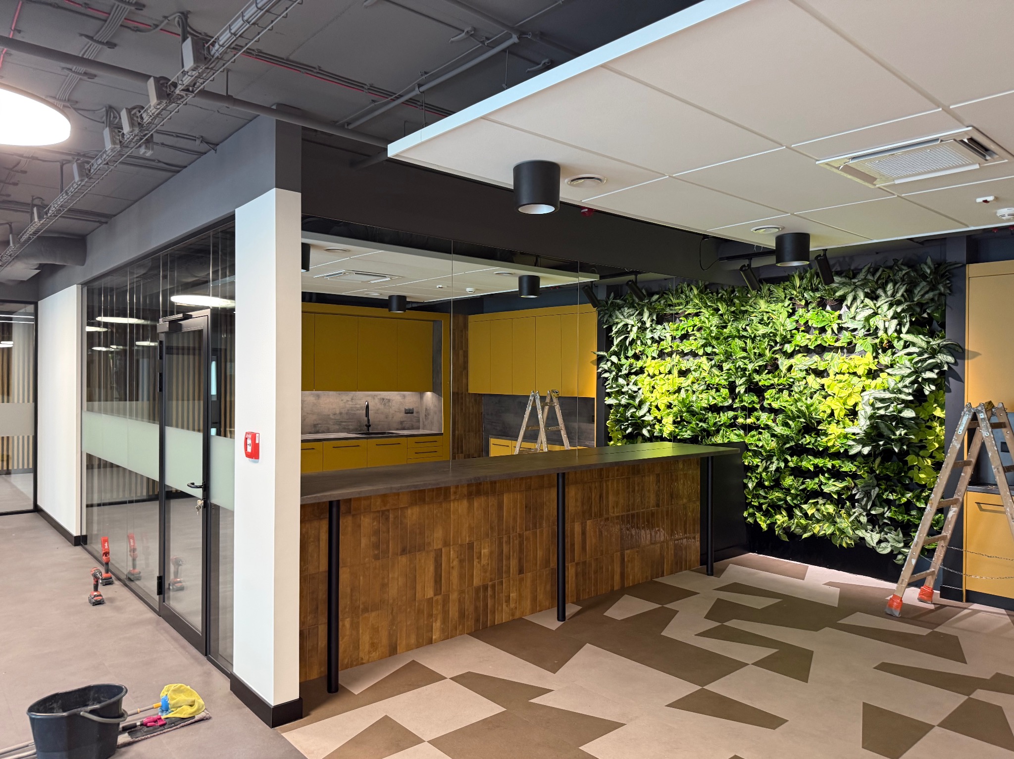 Nowoczesne biuro po remoncie: żółta kuchnia, ściana z roślin, wzorzysta podłoga i drabina. Widoczne narzędzia sugerują świeżo zakończone prace.