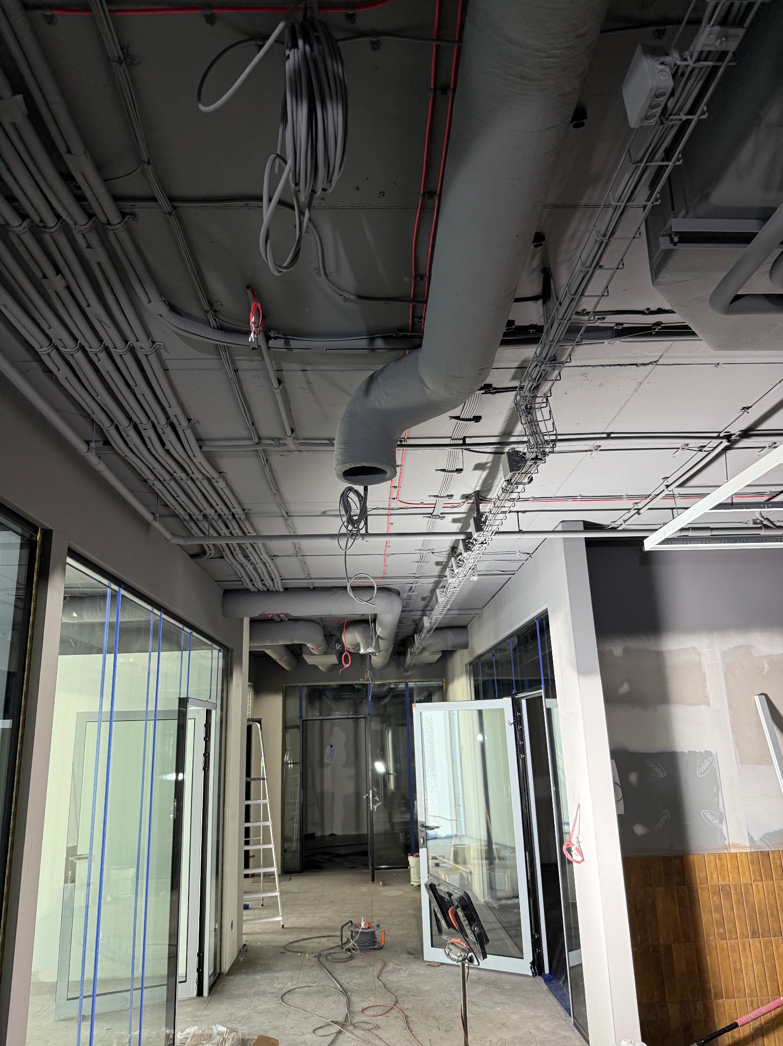 Szare rury wentylacyjne i kable pod sufitem w biurze w trakcie wykańczania. Widoczne szklane ścianki działowe i drabina. Surowy stan, przygotowanie do malowania.