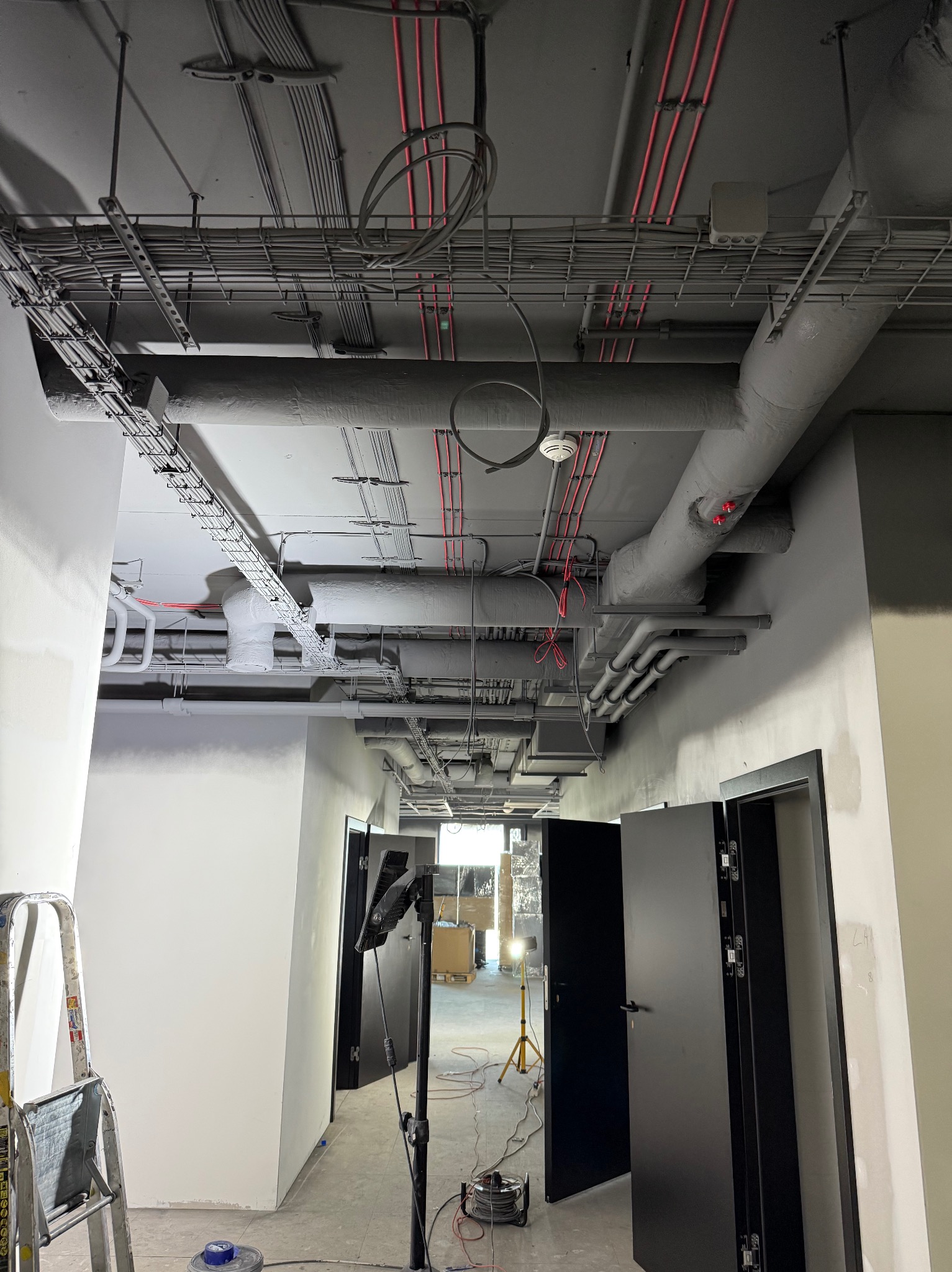 Wnętrze biura w trakcie malowania: szare rury i kable pod sufitem, białe ściany w trakcie malowania, drabina i lampa budowlana. Widoczne otwarte drzwi.