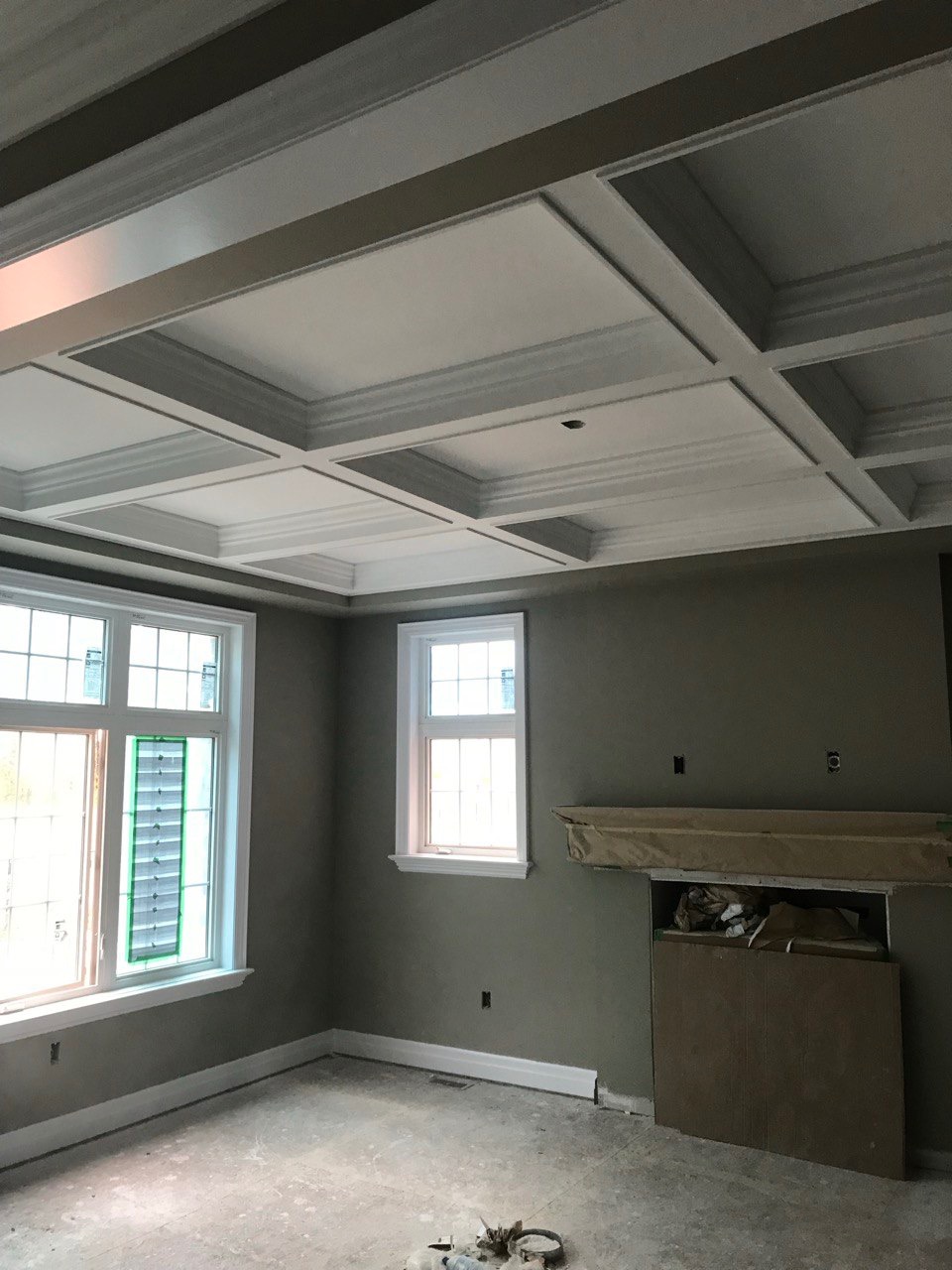 Pokój po szpachlowaniu i malowaniu ścian na szaro, z dekoracyjnym sufitem kasetonowym i dwoma oknami. Podłoga przygotowana do dalszych prac wykończeniowych.