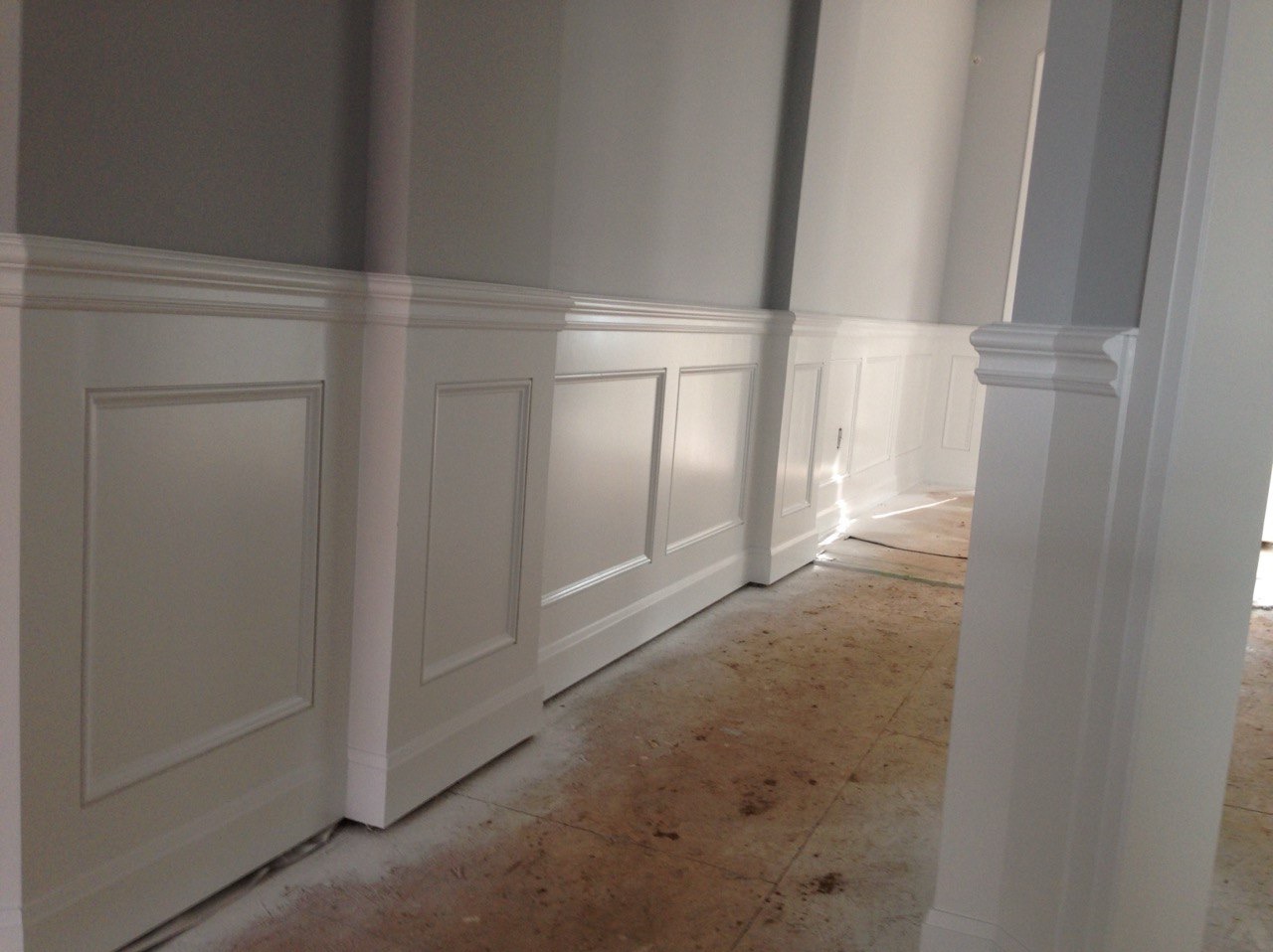 Białe panele ścienne z dekoracyjnymi listwami w jasnym wnętrzu. Ściany pomalowane na szaro, podłoga w trakcie prac wykończeniowych. Klasyczny styl.