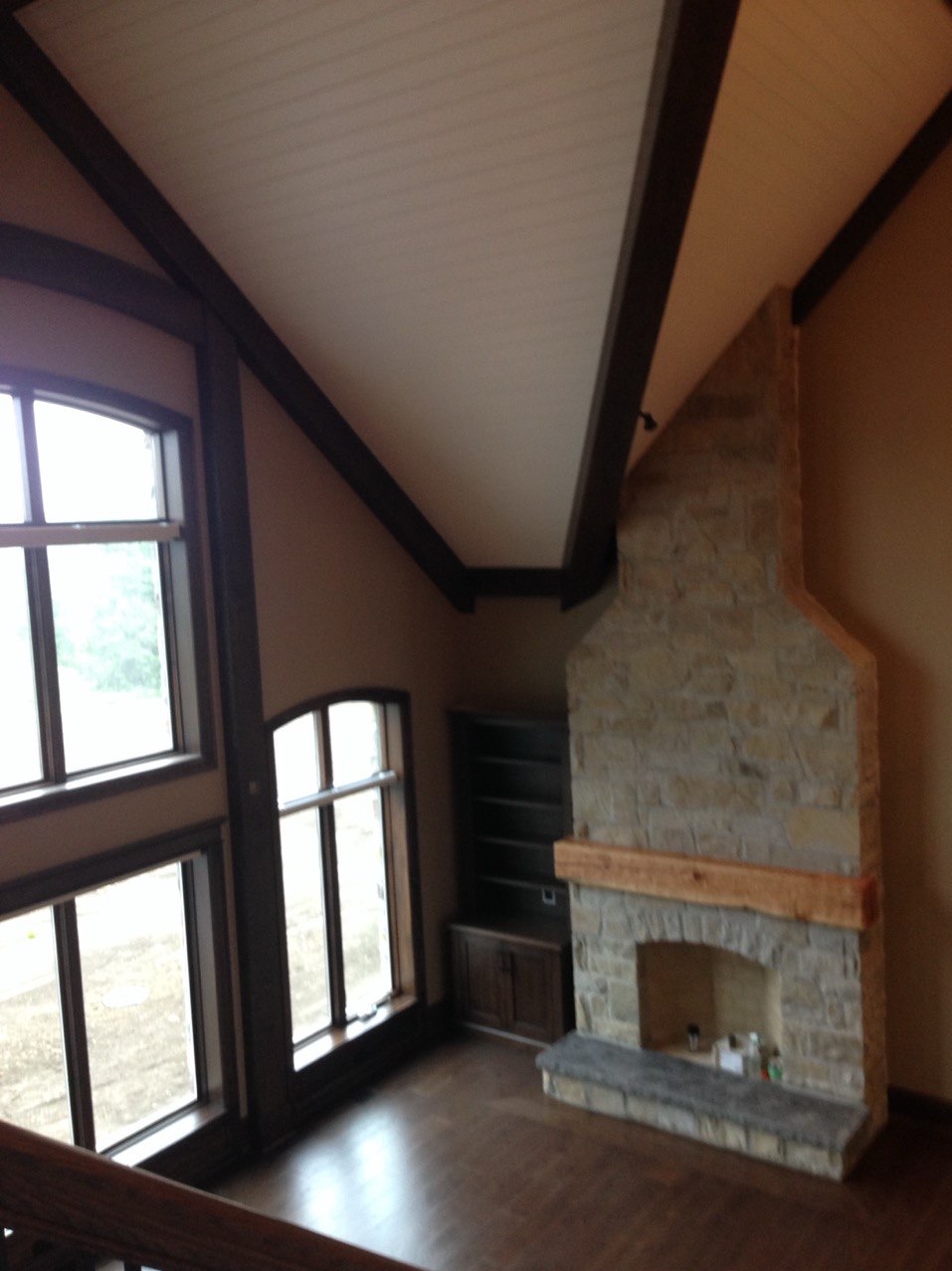 Jasne wnętrze z kamiennym kominkiem i dużymi oknami. Ściany w odcieniach beżu, drewniane belki stropowe. Widok z góry na przestronny salon z parkietem.