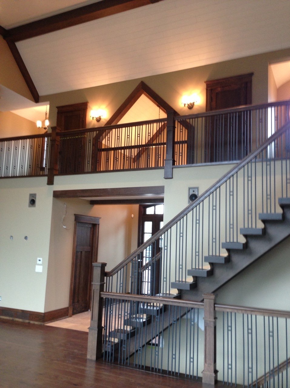 Dwupoziomowe wnętrze z drewnianymi schodami i balustradą. Ściany w odcieniach beżu, drewniane wykończenia sufitu i drzwi. Widoczne oświetlenie i elementy dekoracyjne.