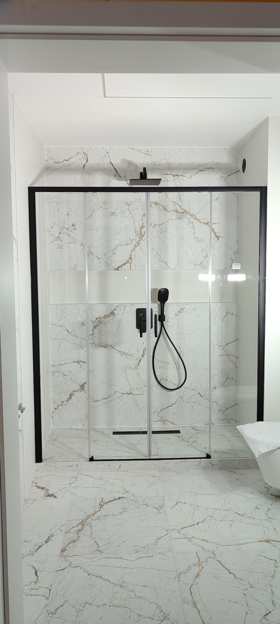 Nowoczesna kabina prysznicowa z czarną ramą i armaturą na tle marmurowych płytek w odcieniach bieli i złota. Minimalistyczny design, eleganckie wykończenie.