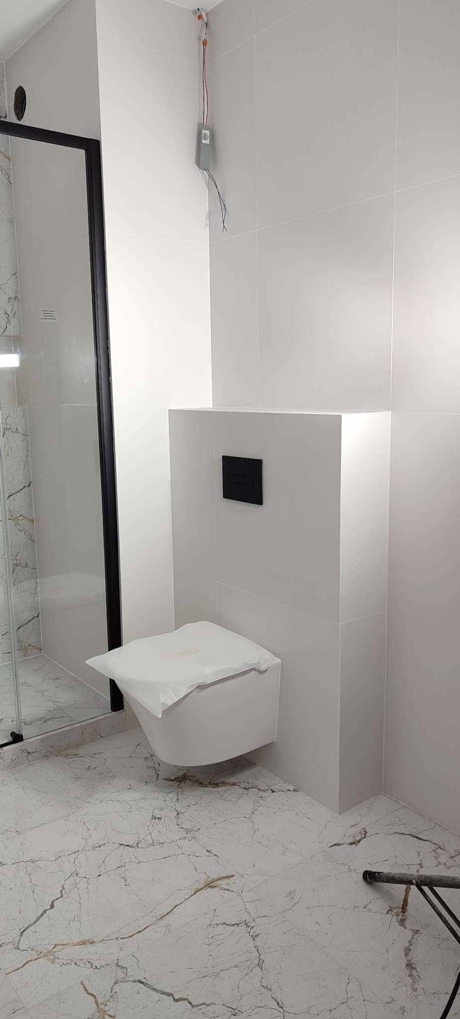 Nowoczesna łazienka z podwieszaną toaletą i kabiną prysznicową. Białe płytki na ścianach i podłodze z marmurowym wzorem. Minimalistyczny design, czarne akcenty.