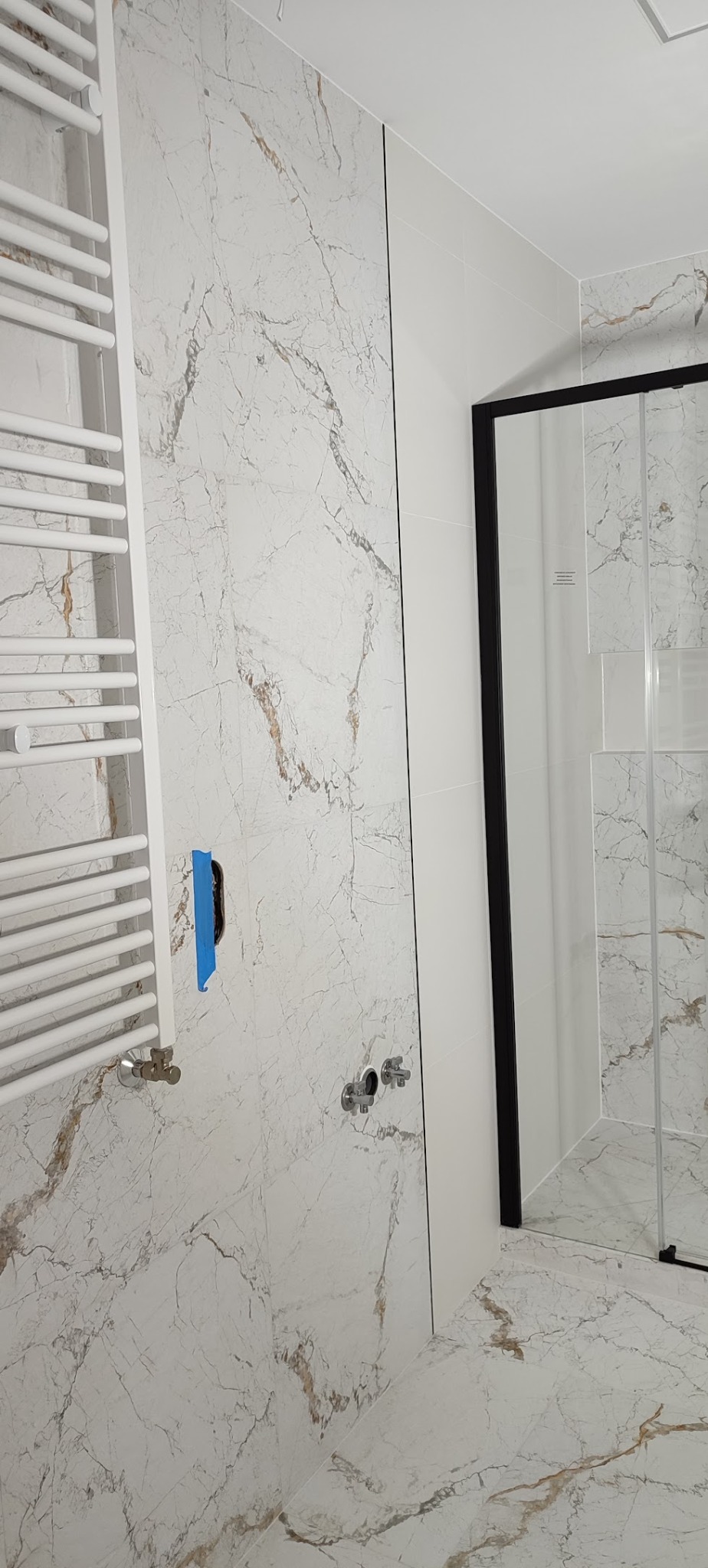 Wykończona łazienka z marmurowymi płytkami, grzejnikiem drabinkowym i kabiną prysznicową z czarną ramą. Częściowo widoczne elementy instalacji.