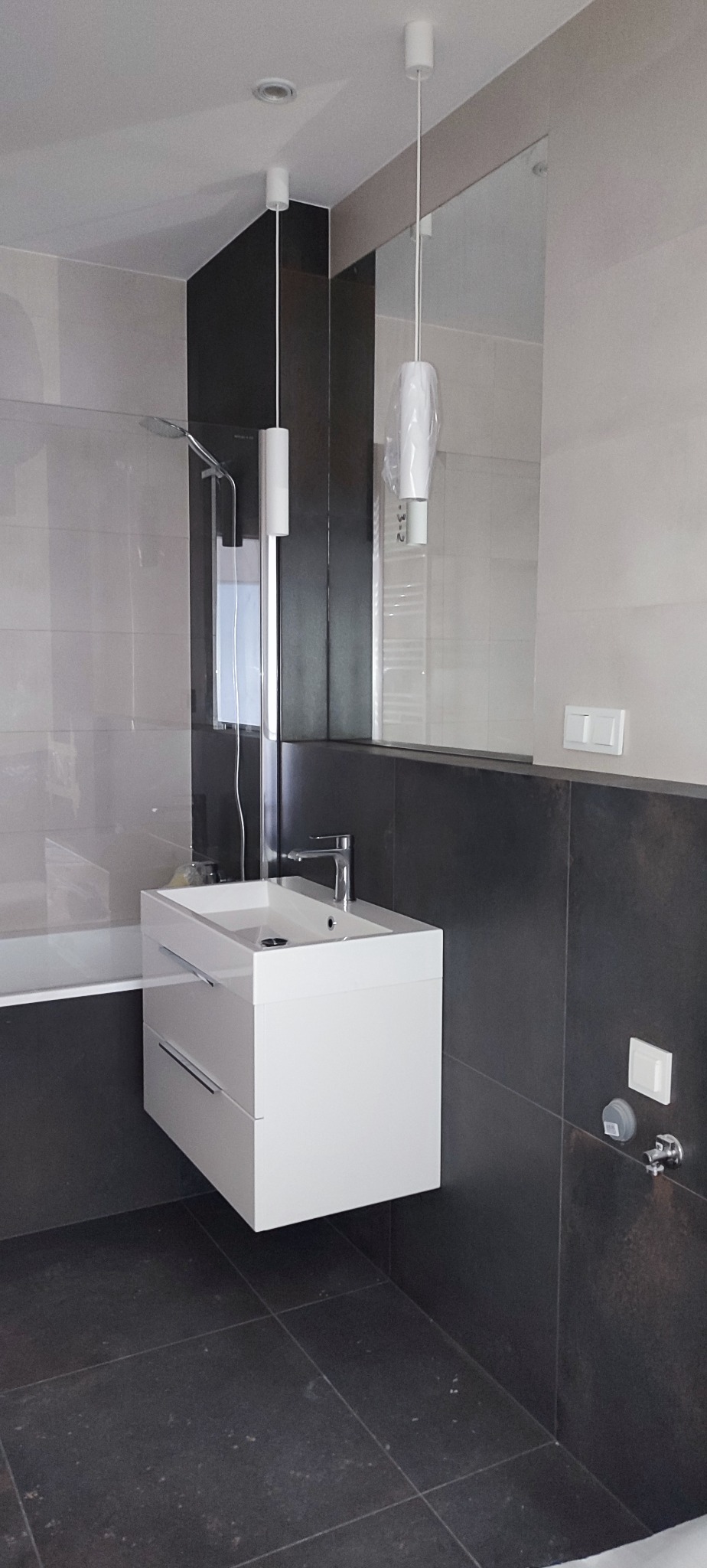 Nowoczesna łazienka z białą szafką pod umywalką, czarnymi płytkami na ścianie i podłodze. Widoczne lustro i lampy wiszące. Kabina prysznicowa w tle.