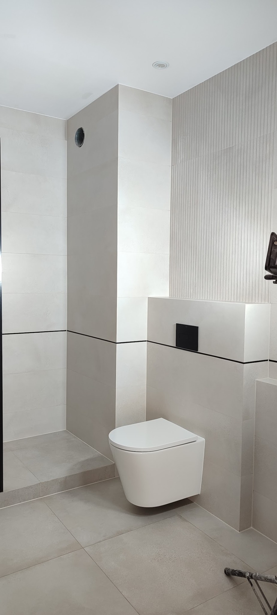 Nowoczesna łazienka z podwieszaną toaletą i beżowymi płytkami, wykończona czarnymi akcentami. Widoczne pionowe żłobienia na ścianie i minimalistyczny design.