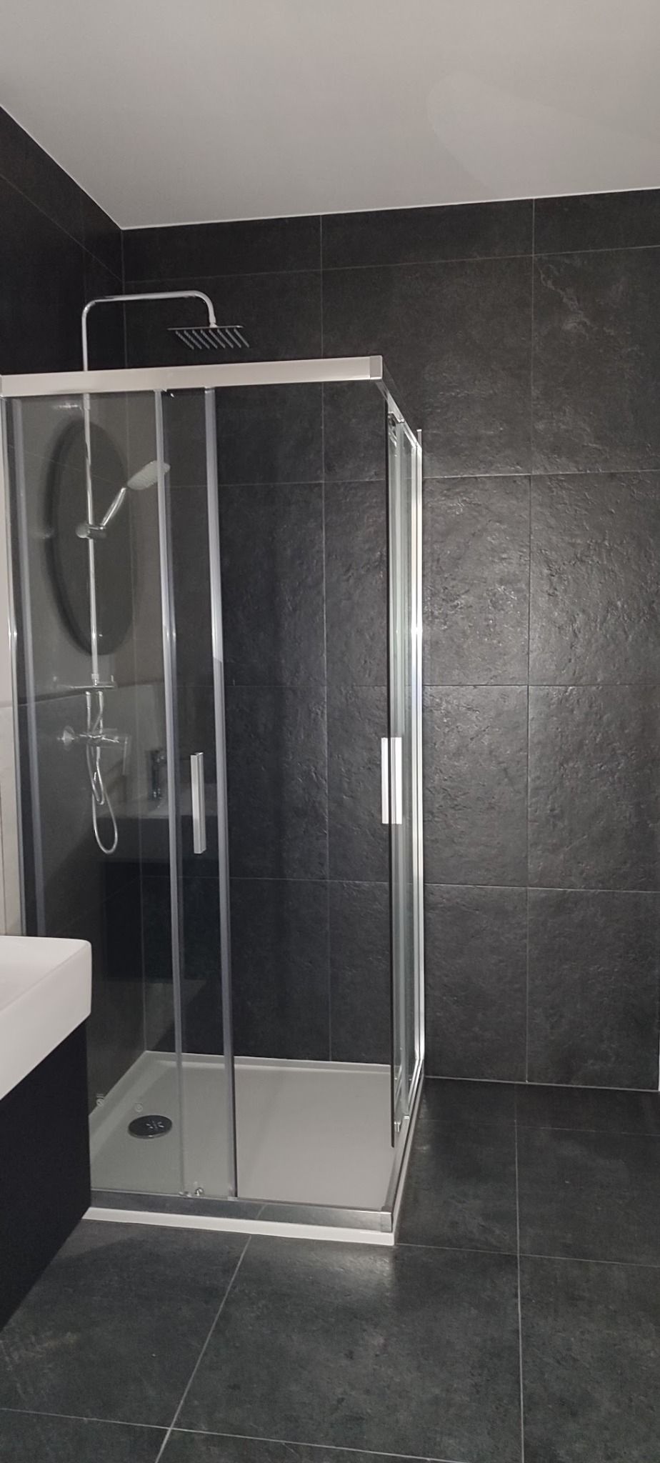 Nowoczesna kabina prysznicowa z deszczownicą, czarne płytki imitujące kamień na ścianach i podłodze, minimalistyczna umywalka z szafką w tle.