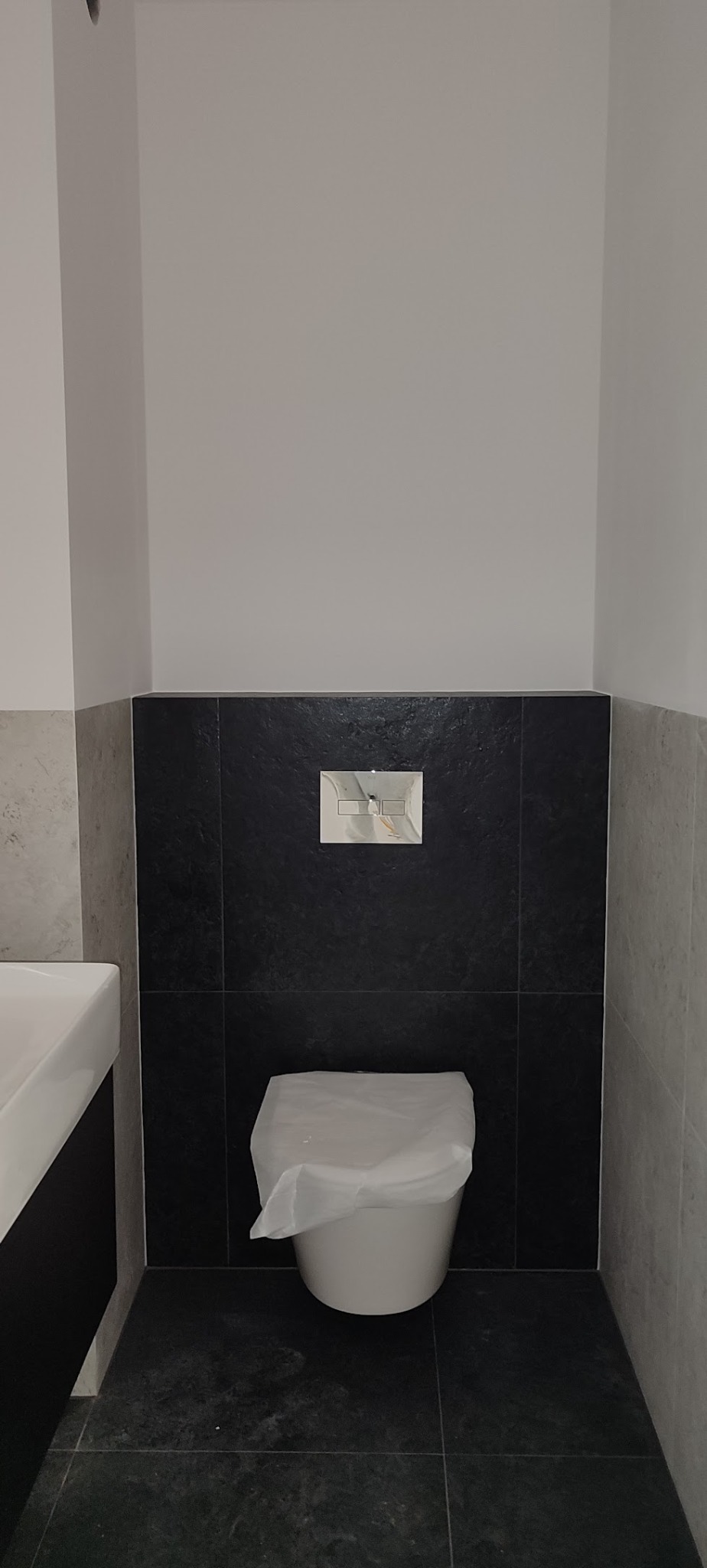 Wykończona łazienka z czarnymi płytkami na ścianie i podłodze, białą miską WC zabezpieczoną folią i chromowanym przyciskiem spłukiwania. Minimalistyczny design.