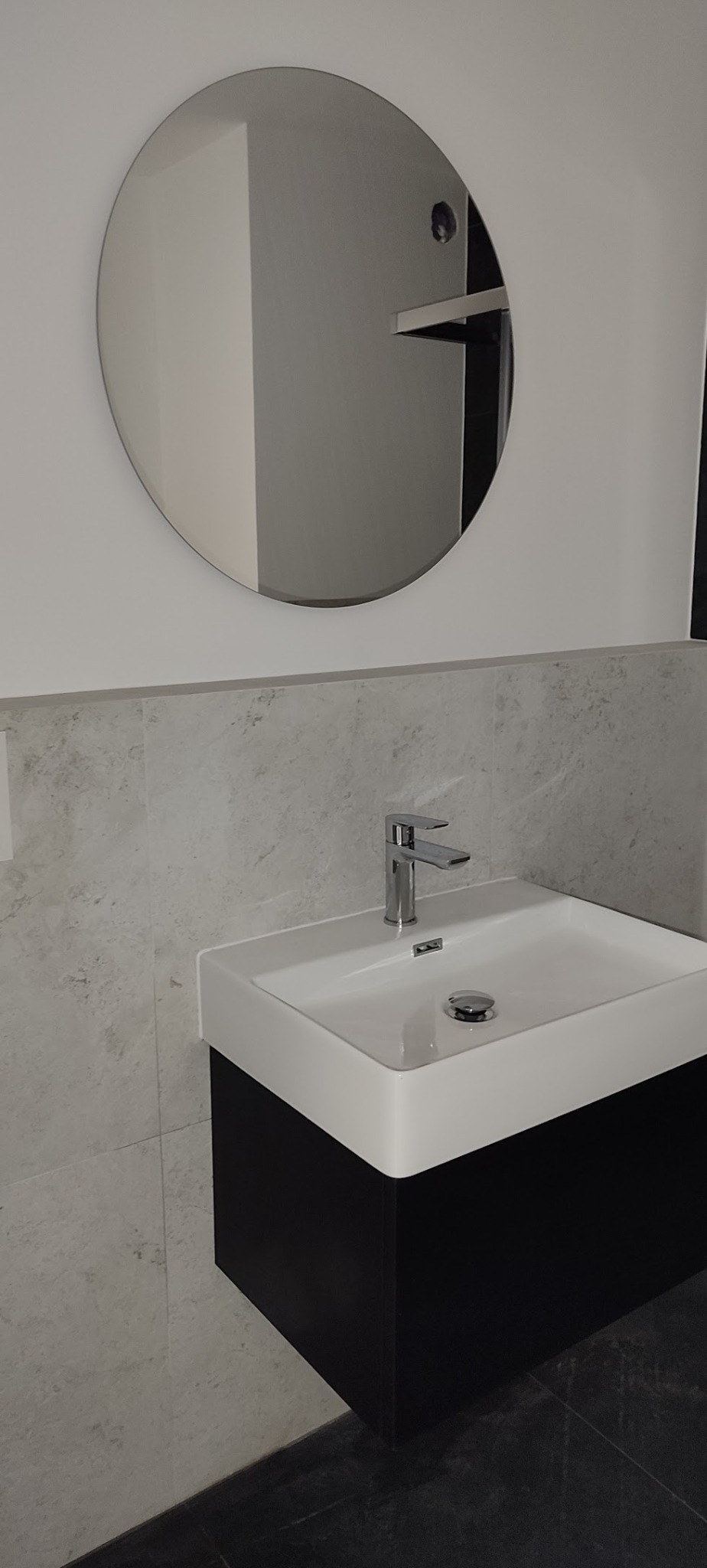 Nowoczesna łazienka: biała umywalka z czarną szafką, chromowana bateria i okrągłe lustro na ścianie z szarymi płytkami imitującymi kamień.