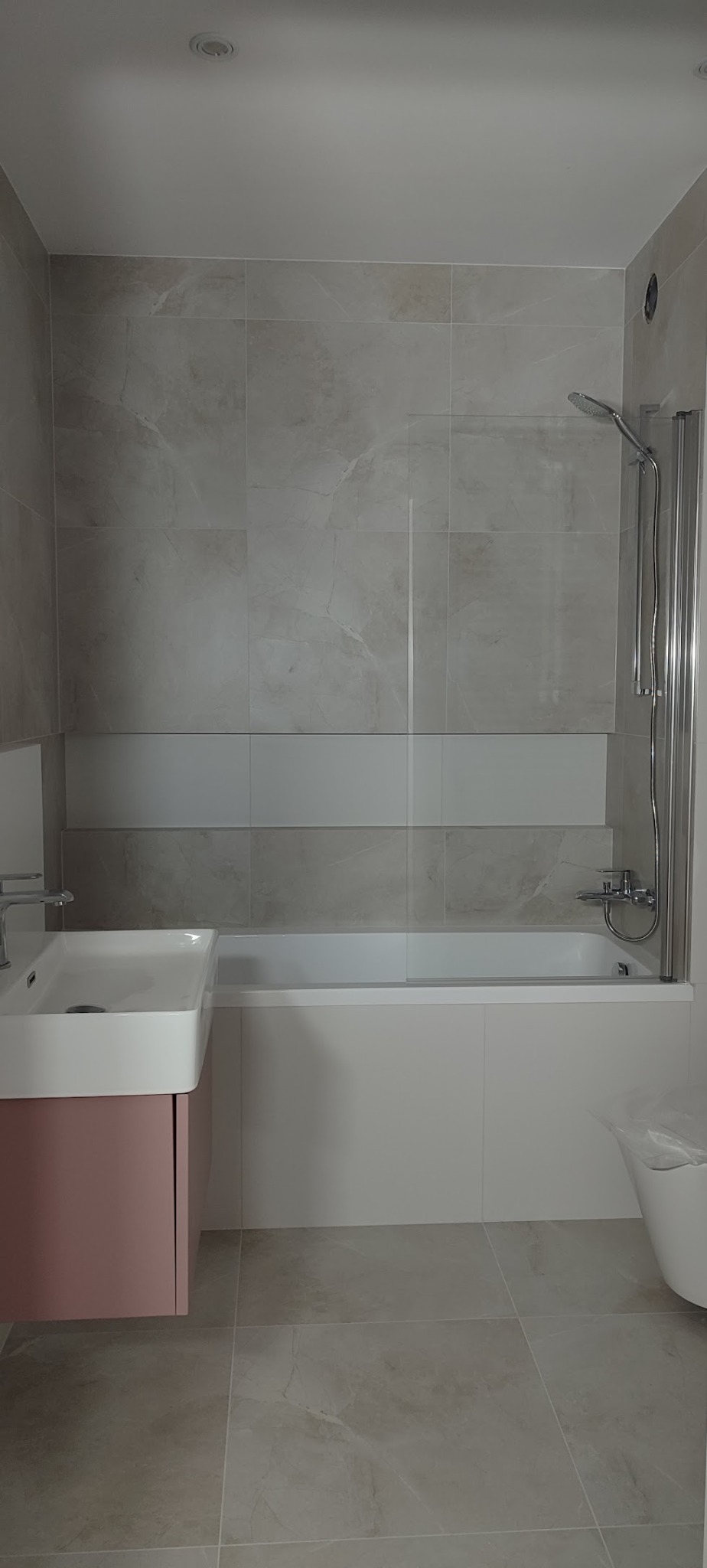 Wykończona łazienka z beżowymi płytkami imitującymi marmur, umywalką na różowej szafce i białą wanną. Kabina prysznicowa ze szklanym parawanem.