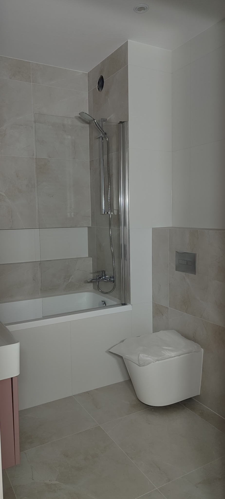 Jasna łazienka po remoncie: beżowe płytki, wanna z parawanem, podwieszana toaleta z osłoną. Minimalistyczny styl, neutralne barwy, widoczne detale.