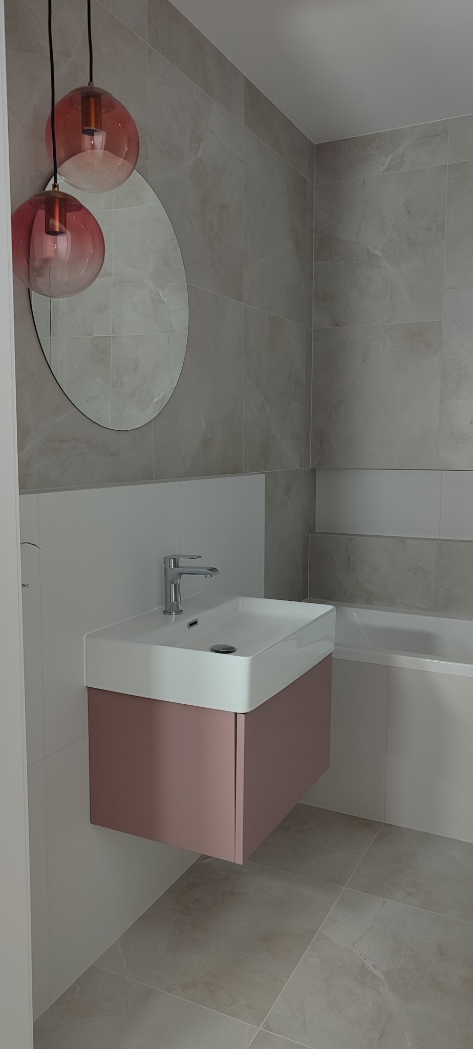 Nowoczesna łazienka z umywalką nablatową na szafce w odcieniach różu, okrągłe lustro i lampy wiszące. Płytki imitujące marmur na ścianach i podłodze.