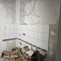 M&S GLOBAL SPÓŁKA Z OGRANICZONĄ ODPOWIEDZIALNOŚCIĄ - Układanie białych płytek w rogu pomieszczenia, widoczne metalowe profile i kartony po płytkach. Instalacja elektryczna na ścianie, etap prac wykończeniowych.